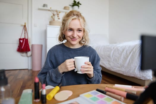 Image of young woman, makeup vlogger, sitting in bedroom with digital camera, drinking tea and talking, creating lifestyle video, social media content.