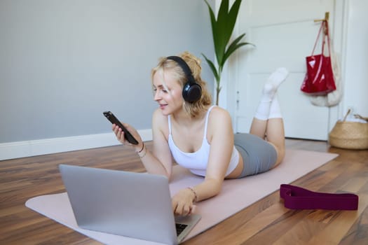 Portrait of young fitness woman, choosing workout on laptop, wearing headphones and using mobile phone exercise app. Sport and lifestyle concept