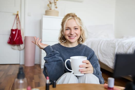 Cheerful woman, beauty blogger, records lifestyle vlog on digital camera, talks casually, tells a story for social media followers, holds cup, drinks tea and sits on floor in her room.