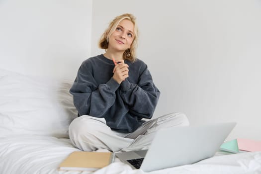 Portrait of cute smiling woman, sitting on a bed, looking dreamy, thinking, using laptop for remote work.