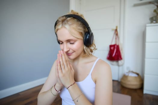 Portrait of young relaxed woman, sits in room in headphones, clasp hands together, meditated, listens to yoga music.