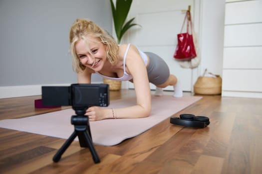 Portrait of young sporty vlogger, fitness instructor standing in plank on rubber yoga mat, recording video of herself doing workout at home.