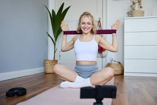 Portrait of young smiling woman, fitness instructor recording video about workout, showing how to exercise at home and use rubber resistance band, sitting on yoga mat.