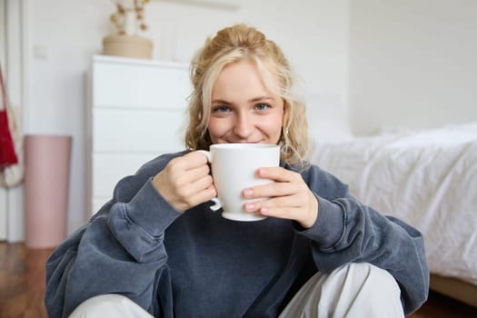 Image of young teenage girl sitting in her bedroom on floor, drinking cup of tea and enjoying day at home, smiling and looking at camera.