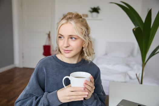 Close up portrait of smiling young blond woman, sits with cup of tea in bedroom, rests at home, enjoys her coffee.