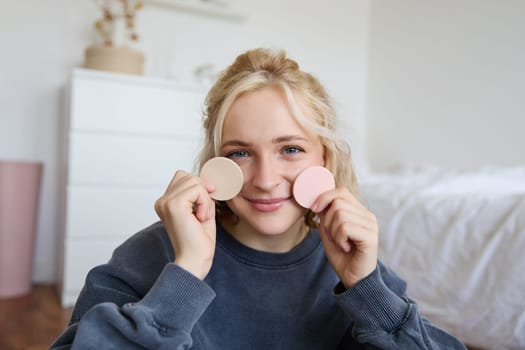 Image of young blond woman, girl records video about makeup, shows skin tone beauty products, sits in a room on floor.
