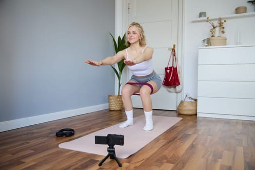 Portrait of young woman, fitness instructor making a video for sport vlog, doing squats on camera, using resistance band, working out at home.