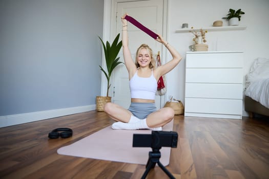 Smiling young blond woman recording video about workout, shooting on digital camera, fitness instructor shows how to use resistance band during exercises. Lifestyle and wellbeing concept