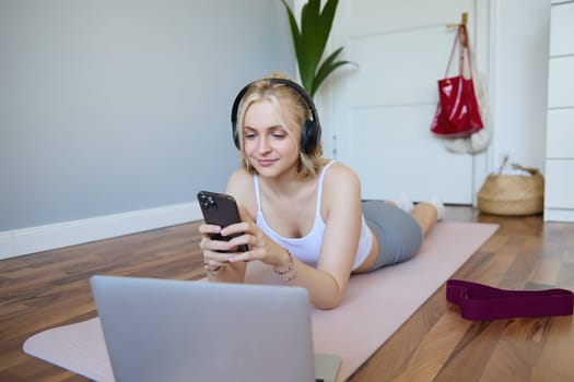 Portrait of young woman workout, watching exercise videos on laptop in headphones, lying on rubber mat with mobile phone and smiling.