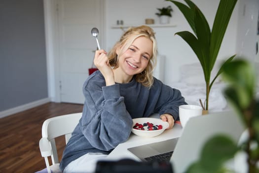 Portrait of smiling blond young woman, eating in front of laptop, watching videos online while having breakfast, enjoying dessert, sitting in bedroom.