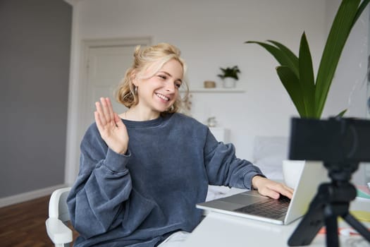 Image of smiling beautiful young woman, chats online on laptop, waves hand, says hello, records video on digital camera, vlogging, creates content for her blog.