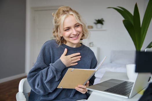 Portrait of smiling, beautiful young blond woman, student working on assignment from home, online learning in her bedroom, talking to video camera, chatting, holding notebook.