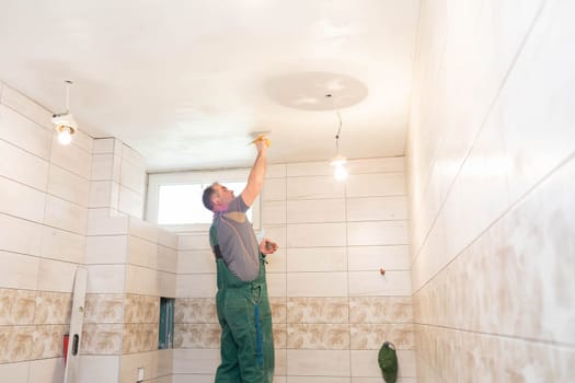 The worker applies plaster on the ceiling of the renovated bathroom. Construction worker in middle age. Green work clothes.