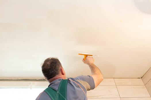 The worker applies plaster on the ceiling of the renovated bathroom. Construction worker in middle age. Green work clothes.