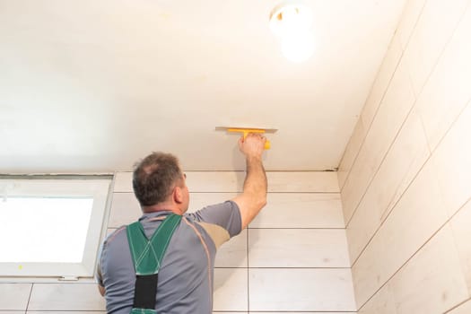 The worker applies plaster on the ceiling of the renovated bathroom. Construction worker in middle age. Green work clothes.