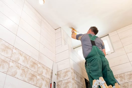 The worker applies plaster on the ceiling of the renovated bathroom. Construction worker in middle age. Green work clothes.
