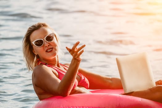 Woman works on laptop in sea. Freelancer, young blond woman in sunglases floating on an inflatable big pink donut with a laptop in the sea at sunset. Freelance, travel and holidays concept