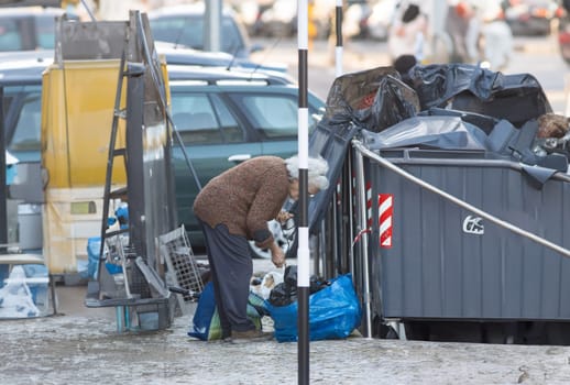 An old woman is sorting through trash in a dumpster. The scene is messy and disorganized, with trash scattered around the area. The woman is working hard to clean up the area