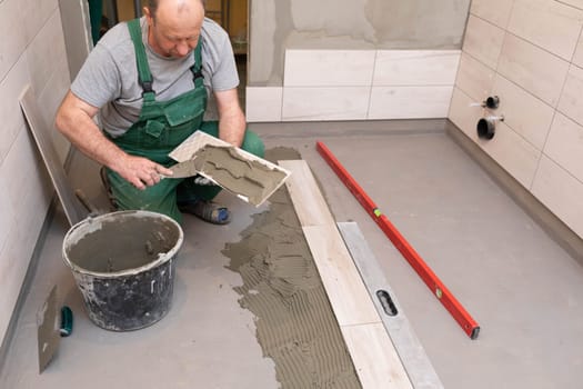 A construction worker lubricates the bottom of the ceramic tile with a metal trowel with special glue.