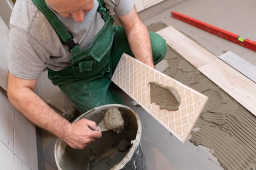 A construction worker lubricates the bottom of the ceramic tile with a metal trowel with special glue.