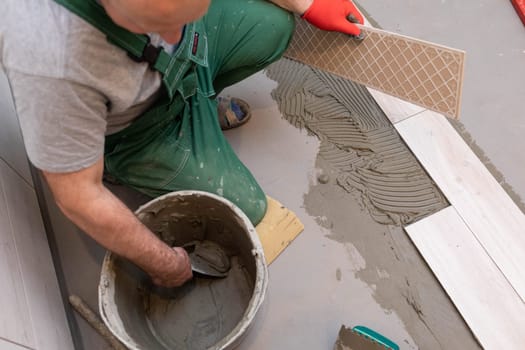 A construction worker lubricates the bottom of the ceramic tile with a metal trowel with special glue.