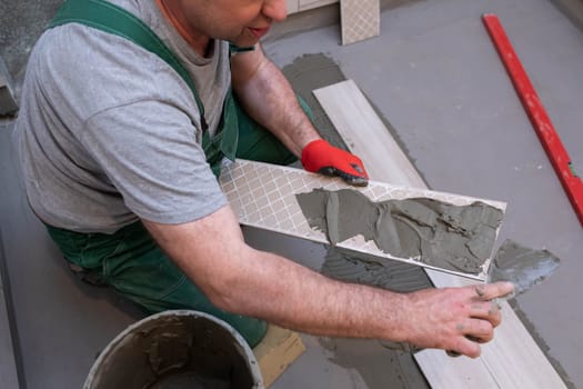 A construction worker lubricates the bottom of the ceramic tile with a metal trowel with special glue.