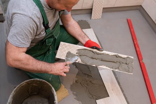 A construction worker lubricates the bottom of the ceramic tile with a metal trowel with special glue.