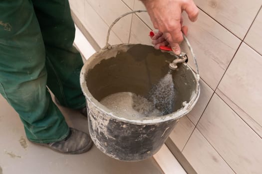 Construction worker. Unscrewed tap. Tap water flows into the bucket. During renovation. Tiles on the bathroom wall.