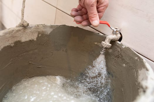 Construction worker. Unscrewed tap. Tap water flows into the bucket. During renovation. Tiles on the bathroom wall.