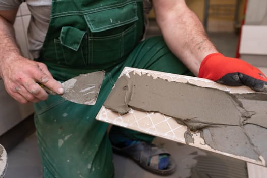 A construction worker lubricates the bottom of the ceramic tile with a metal trowel with special glue.