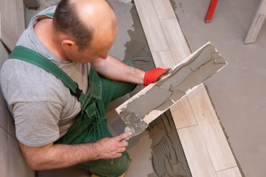 A construction worker lubricates the bottom of the ceramic tile with a metal trowel with special glue.