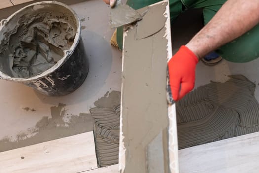 A construction worker lubricates the bottom of the ceramic tile with a metal trowel with special glue.