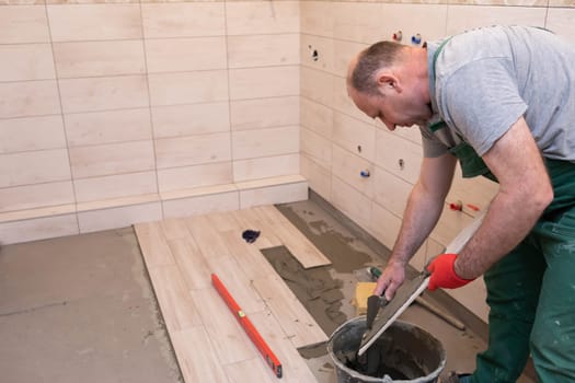 A construction worker lubricates the bottom of the ceramic tile with a metal trowel with special glue.