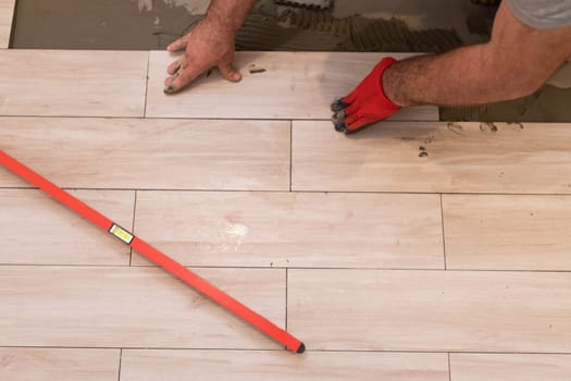 He presses the ceramic tile against the floor with his hand. Protective glove on hand. Red spirit level. A professional construction worker.