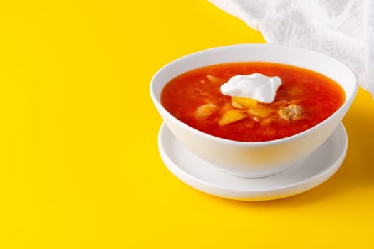 Red borscht in bowl on a yellow background, traditional dish that is cooked with broth, with copy space for text.