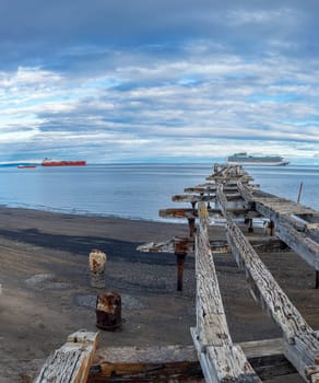 Aged wooden pier extends into serene blue sea with overcast skies and ships on the horizon.