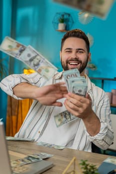 Cheerful rich Caucasian young freelancer man working on laptop and wasting throwing money dollar cash more tips earnings big profit success win lottery shopping. Guy sitting at home office. Vertical