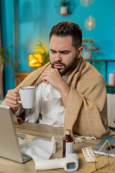 Sick ill young businessman suffering from cold flu or allergy working on laptop at home office. Manager freelancer man wrapped in blanket blows sneezes wipes snot into napkin. Coronavirus quarantine.