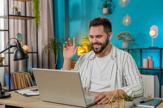 Caucasian young freelancer man looking at laptop screen, making video webcam conference call with friends or family enjoying pleasant conversation. Guy sits at home office table workplace. Lifestyle
