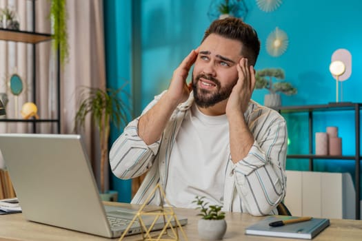 Tired ill businessman suffering from headache problem tension and migraine, stress at home office desk workplace. Bearded freelancer broker worker man works on laptop notebook. Overworking. Lifestyle