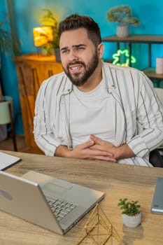 Disappointed freelancer man feeling overeating during work process on laptop in home office. Upset businessman freelancer guy man touching stomach and experiencing discomfort and sharp pain. Vertical
