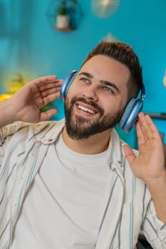 Happy cheerful Caucasian young man sits at home office desk listening to energetic disco music through wireless headphones. Freelancer guy relaxing taking a break with laptop on table. Vertical