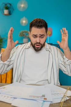 Frustrated upset young business man works at office desk looking at bills paperwork reports documents gets angry, frustrated, stress bankruptcy financial problem. Stresses freelancer at home. Vertical