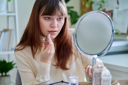 Young woman with cosmetic products on table, doing makeup looking in mirror at home, painting lips with lipstick. Cosmetics, fashion, youth, lifestyle, beauty concept