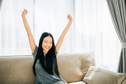 Happy woman on sofa raises arms stretches after sitting for long time. Lifestyle of a cheerful businesswoman in a luxury living room. Wellbeing joy and carefree relaxation.