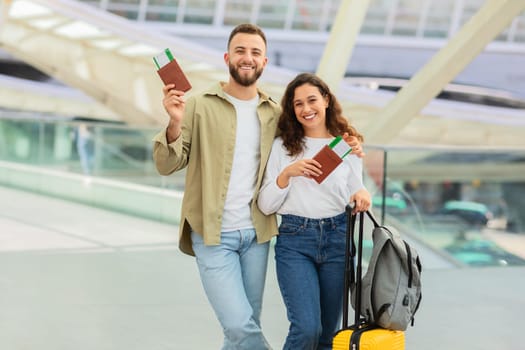 Happy Loving Young Spouses Embracing In Airport Terminal While Waiting Flight, Romantic Millennial Couple With Suitcase, Passport And Tickets Standing In Lounge Zone, Ready For Honeymoon Trip