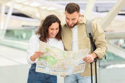 Content young couple reading city map together while standing in airport terminal, happy millennial man and woman planning vacation travel, checking journey route and smiling, free space