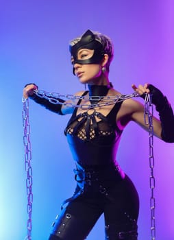 a sexy girl in the erotic image of a catwoman in leather belts and a mask poses against the background of a copy paste in a beautiful neon background with a metal chain in her hands