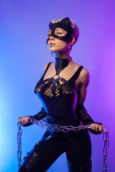 a sexy girl in the erotic image of a catwoman in leather belts and a mask poses against the background of a copy paste in a beautiful neon background with a metal chain in her hands