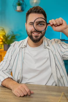 Caucasian young freelancer man holding magnifying glass near face, looking into camera with big zoomed funny eye, searching, analyzing. Bearded guy sitting at home office table workplace. Vertical